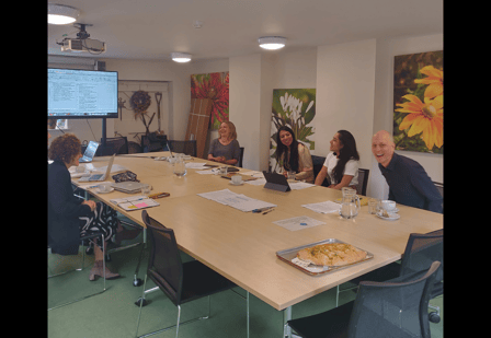 Five people sat around a table facing a screen with a presentation displayed