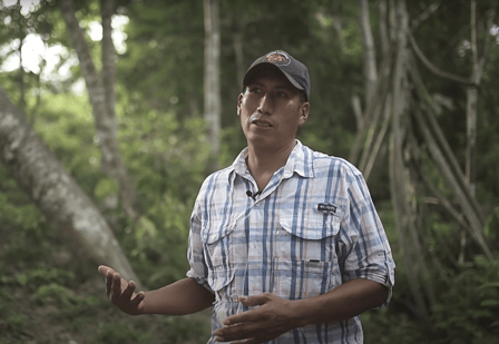 Man in checked shirt and baseball cap speaking to camera. Dense forest in background.