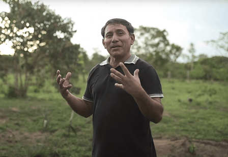 Man in black polo shirt speaking to camera. Fields with trees in background.