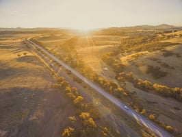 Aerial shot of a road with a few vehicles. Sun setting at the horizon. Green space on either side of the road. 