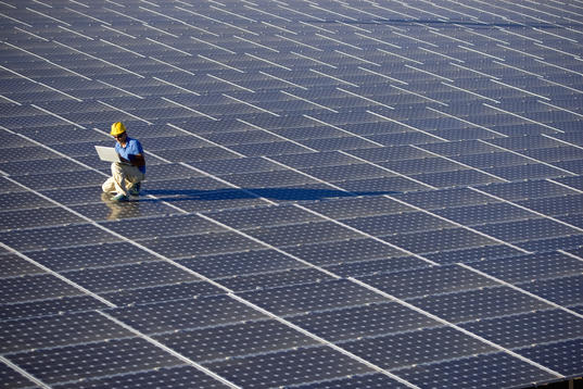 Man sitting in a hard hat and working on his laptop against the backdrop of solar panels