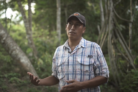 Man in checked shirt and baseball cap speaking to camera. Dense forest in background. 