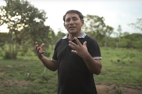 Man in black polo shirt speaking to camera. Fields with trees in background.