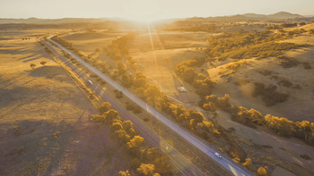 Aerial shot of a road with a few vehicles. Sun setting at the horizon. Green space on either side of the road. 